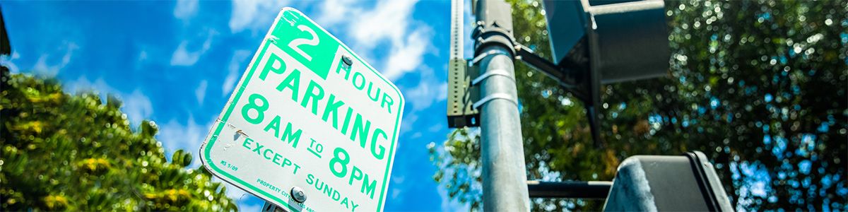 A green and white 2-hour parking sign against a blue sky, a metal street light pole, and green tree tops.