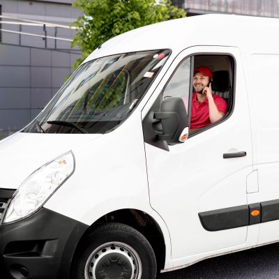 A man is in a white delivery van is wearing a red uniform, smiling, and talking into a cell phone.