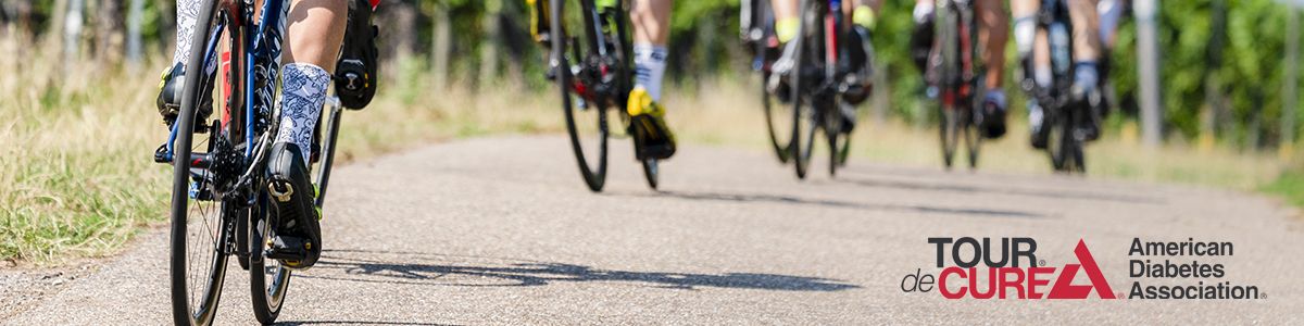 A shot of a team of bicycle riders wearing colorful socks, riding down the road. To the lover right is the logo for the Tour De Cure - American Diabetes Association 